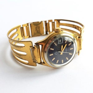 Könnte beinhalten: Eine goldfarbene Armbanduhr mit schwarzem Zifferblatt und einem goldenen Metallband. Das Zifferblatt der Uhr hat ein Datumsfenster und den Text "SLAVICA" darauf gedruckt.