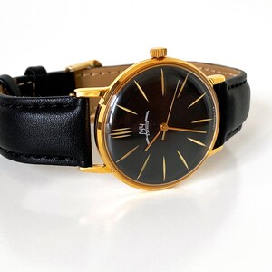 May include: A gold-tone wristwatch with a black face and gold hands. The watch has a black leather band.