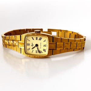 K&ouml;nnte beinhalten: Eine goldfarbene Armbanduhr mit quadratischem Zifferblatt und einem goldfarbenen Metallband. Das Zifferblatt der Uhr hat r&ouml;mische Ziffern als Stundenmarkierungen und einen schwarzen Sekundenzeiger.