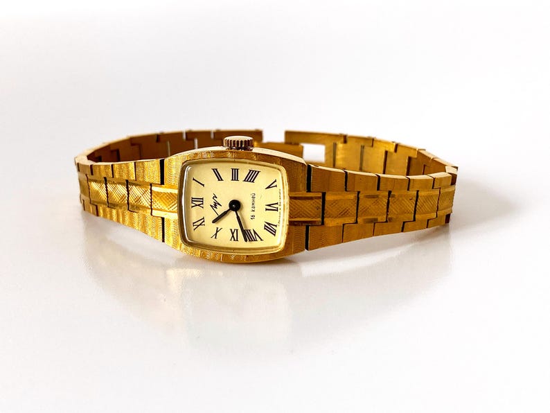 K&ouml;nnte beinhalten: Eine goldfarbene Armbanduhr mit quadratischem Zifferblatt und einem goldfarbenen Metallband. Das Zifferblatt der Uhr hat r&ouml;mische Ziffern als Stundenmarkierungen und einen schwarzen Sekundenzeiger.