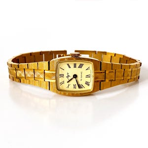 K&ouml;nnte beinhalten: Eine goldfarbene Armbanduhr mit quadratischem Zifferblatt und r&ouml;mischen Ziffern f&uuml;r die Stunden. Die Uhr hat ein goldfarbenes Metallband mit einem strukturierten Muster.