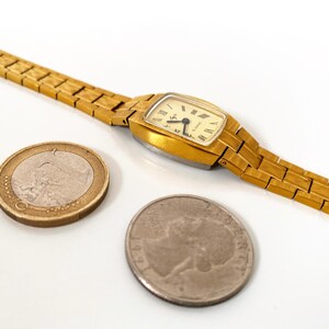 K&ouml;nnte beinhalten: Eine goldfarbene Armbanduhr mit quadratischem Zifferblatt und einem goldfarbenen Metallband. Die Uhr wird neben einer Euro-M&uuml;nze und einer US-Viertel-Dollar-M&uuml;nze gezeigt.