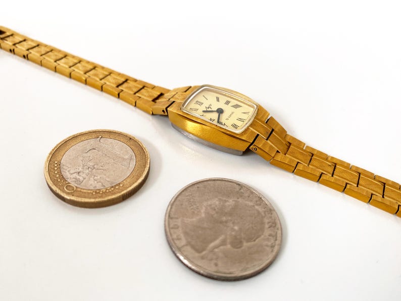 K&ouml;nnte beinhalten: Eine goldfarbene Armbanduhr mit quadratischem Zifferblatt und einem goldfarbenen Metallband. Die Uhr wird neben einer Euro-M&uuml;nze und einer US-Viertel-Dollar-M&uuml;nze gezeigt.