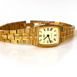 K&ouml;nnte beinhalten: Eine goldfarbene Armbanduhr mit quadratischem Zifferblatt und einem goldfarbenen Metallband. Das Zifferblatt der Uhr hat r&ouml;mische Ziffern als Stundenmarkierungen und einen schwarzen Sekundenzeiger. Auf dem Zifferblatt der Uhr steht auch der Text "Chronotech".
