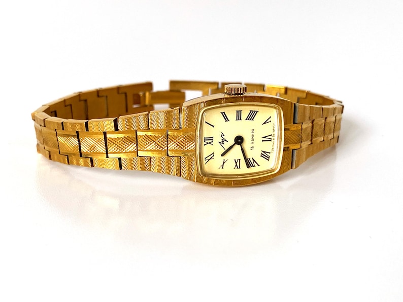 K&ouml;nnte beinhalten: Eine goldfarbene Armbanduhr mit quadratischem Zifferblatt und einem goldfarbenen Metallband. Das Zifferblatt der Uhr hat r&ouml;mische Ziffern als Stundenmarkierungen und einen schwarzen Sekundenzeiger. Auf dem Zifferblatt der Uhr steht auch der Text "Chronotech".