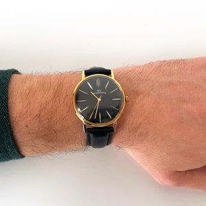 May include: A gold-tone wristwatch with a black face and black leather band. The watch face has gold-tone hands and Roman numeral hour markers.