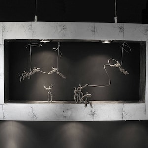 May include: A white framed shadow box with a black background. Inside the box, there are several wire sculptures of people hanging from the ceiling. The sculptures are made of thin wire and are very detailed.
