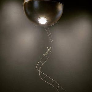 Puede incluir: Una luminaria esférica de color marrón oscuro cuelga del techo con una sola bombilla en su interior. Una fina escalera de alambre plateado se extiende hacia abajo desde la luminaria, con una pequeña figura plateada que sube por la escalera.