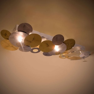 May include: A modern ceiling light fixture with multiple circular metal shades in silver and gold tones. The light fixture is suspended from the ceiling and has a contemporary design.
