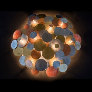 Puede incluir: Lámpara de pared decorativa compuesta por discos metálicos circulares superpuestos en varios colores, incluyendo dorado, plateado, cobre y bronce. La lámpara tiene varias luces pequeñas que iluminan los discos, creando un brillo cálido. El diseño es moderno y artístico.