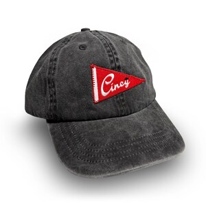 May include: A dark gray baseball cap with a curved brim. A red pennant patch with white trim and the word "Cincy" in cursive is sewn on the front. The cap has a six-panel design and an adjustable strap.
