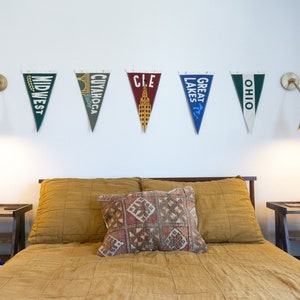 May include: A bedroom with a bed with a yellow quilted bedspread and a patterned pillow. There are two nightstands with lamps on either side of the bed. There are six pennants hanging on the wall above the bed, each with a different city or region name: "Midwest", "Cuyahoga", "CLE", "Great Lakes", "Ohio".