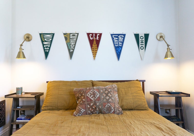 May include: A bedroom with a bed with a yellow quilted bedspread and a patterned pillow. There are two nightstands with lamps on either side of the bed. There are six pennants hanging on the wall above the bed, each with a different city or region name: "Midwest", "Cuyahoga", "CLE", "Great Lakes", "Ohio".