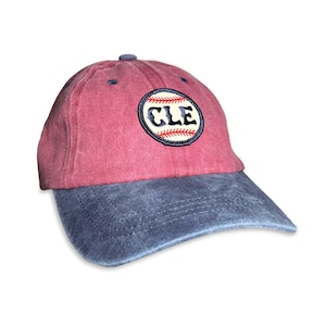May include: A baseball cap with a faded red crown and a dark gray brim. The front features a circular embroidered patch displaying a white baseball graphic with red stitching and the dark blue text CLE centered in the middle of the baseball design.