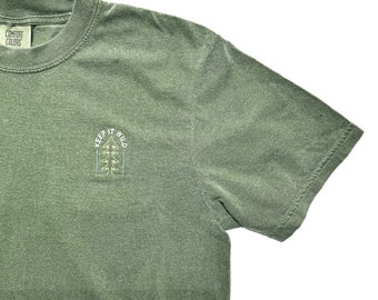May include: A green short-sleeved t-shirt with a small embroidered design on the left chest. The design features a pine tree with the text "Keep it Wild" above it.