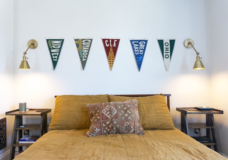 May include: A bedroom with a bed with a yellow duvet and a patterned pillow. There are two nightstands with lamps on either side of the bed. There are six pennants hanging on the wall above the bed, each with a different city or state name on it. The pennants are: "Midwest", "Cuyahoga", "CLE", "Great Lakes", "Ohio".
