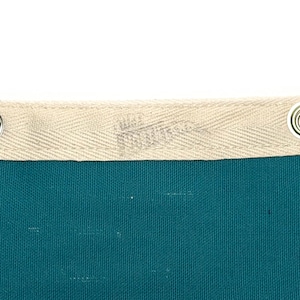 May include: Close-up of a teal fabric with a cream-colored border. The border has a woven texture and metal grommets. The fabric appears to be a banner or flag, with a subtle logo visible on the border.