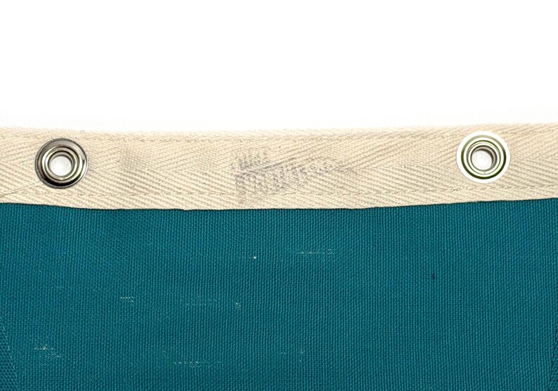 May include: Close-up of a teal fabric with a cream-colored border. The border has a woven texture and metal grommets. The fabric appears to be a banner or flag, with a subtle logo visible on the border.