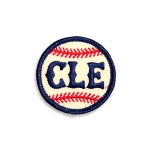 May include: A circular embroidered patch featuring a cream-colored background with red baseball-style stitching at the top and bottom. The letters CLE are stitched in dark navy blue in the center, surrounded by a thick dark navy blue border.