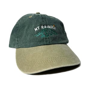 May include: A green and khaki baseball cap with an embroidered image of Mount Rainier and the text "MT RAINIER".