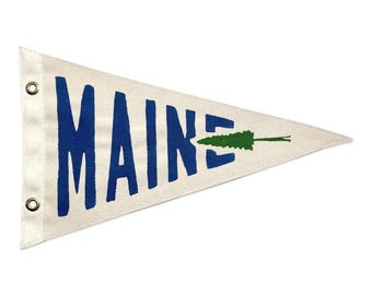 May include: A cream-coloured pennant flag with the word "MAINE" in bold blue letters. A green tree graphic is on the right side. The flag has a white border with metal eyelets for hanging. The pennant is triangular in shape.