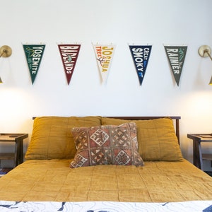 May include: A bedroom scene featuring a bed with mustard-colored bedding and a patterned pillow. Above the bed, five pennant flags with national park names are displayed. Two brass sconces flank the flags. Two wooden nightstands are on either side of the bed.