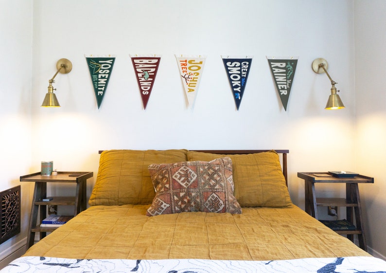 May include: A bedroom scene featuring a bed with mustard-colored bedding and a patterned pillow. Above the bed, five pennant flags with national park names are displayed. Two brass sconces flank the flags. Two wooden nightstands are on either side of the bed.