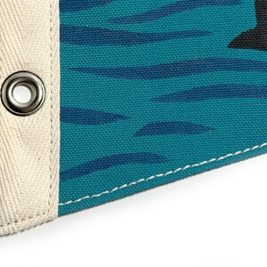 May include: Close-up of a teal canvas pennant flag with a black graphic and dark blue horizontal stripes. The flag has a cream-colored canvas border with a metal grommet and white stitching.