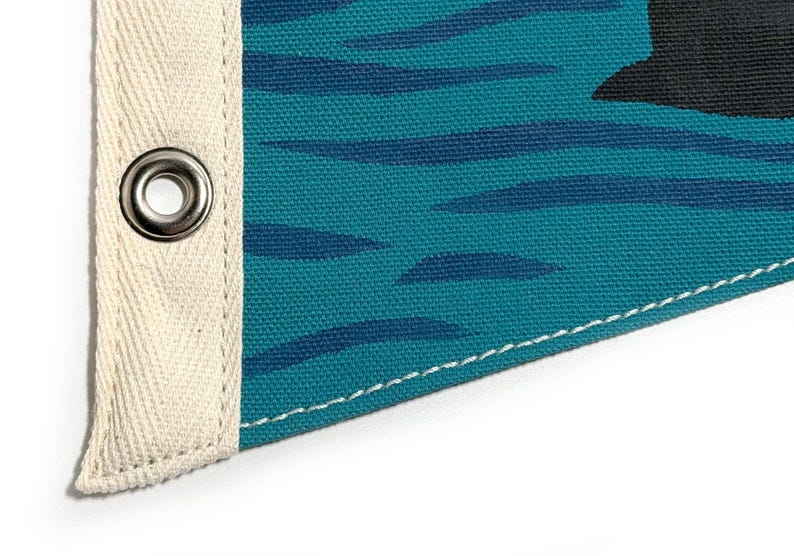 May include: Close-up of a teal canvas pennant flag with a black graphic and dark blue horizontal stripes. The flag has a cream-colored canvas border with a metal grommet and white stitching.