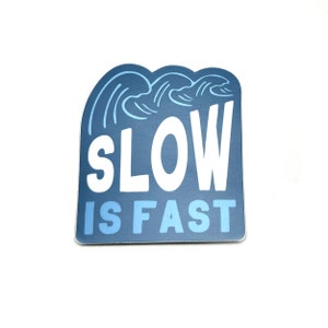 May include: A sticker with a blue background and white text that reads "SLOW IS FAST." The top of the sticker features a stylized wave design in shades of blue. The sticker has a rounded rectangular shape.