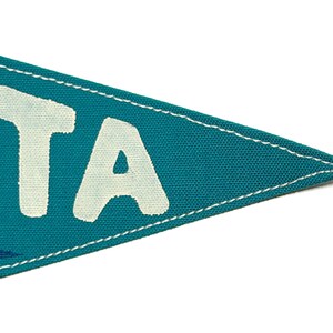 May include: Teal pennant flag with white text reading "OTA". The flag has a white stitched border and a blue wave design. The pennant is made of fabric and is set against a white background.