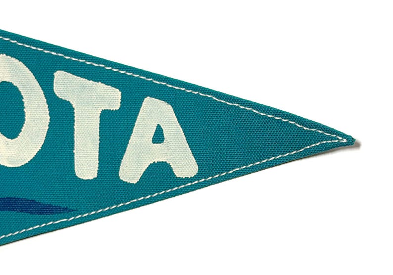 May include: Teal pennant flag with white text reading "OTA". The flag has a white stitched border and a blue wave design. The pennant is made of fabric and is set against a white background.