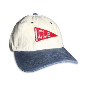 May include: A baseball cap with an off-white crown and a distressed blue denim brim. The front features an embroidered red pennant flag patch with the white letters CLE stitched inside. The cap includes small dark blue eyelets on the crown.