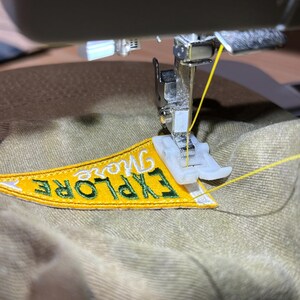 May include: A sewing machine is stitching a yellow and green embroidered patch onto a beige fabric. The patch reads "Explore More".