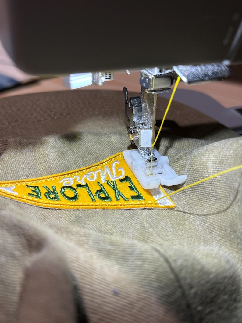 May include: A sewing machine is stitching a yellow and green embroidered patch onto a beige fabric. The patch reads "Explore More".