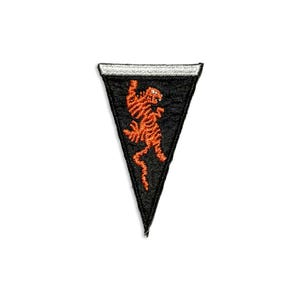 May include: A triangular black embroidered patch featuring a stylized orange tiger figure in the center. The patch has a white border along the top edge and a black stitched border around the remaining sides, creating a pennant-like shape.