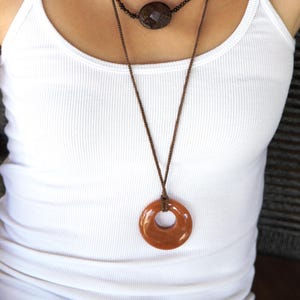 May include: Two necklaces with brown cords. The top necklace has a dark brown, faceted, circular pendant. The bottom necklace features a large, orange-brown, donut-shaped pendant. The necklaces are worn over a white tank top.