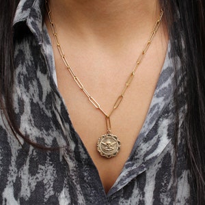 May include: A gold chain necklace with a gold pendant featuring an angel design. The necklace is worn by a person with long dark hair and a gray and white patterned shirt.