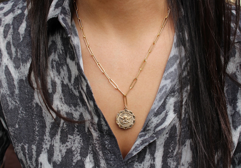 May include: A gold chain necklace with a gold pendant featuring an angel design. The necklace is worn by a person with long dark hair and a gray and white patterned shirt.