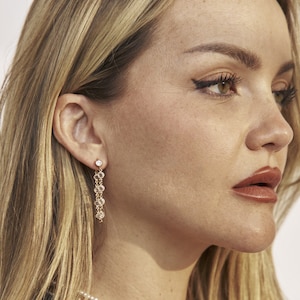 May include: A close-up of a woman's face, showing her right ear with a gold chain earring with small clear stones. The earring dangles from the earlobe and has a single larger clear stone at the top.