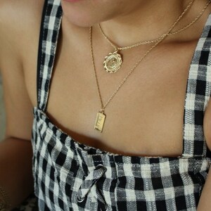 May include: A woman wearing a black and white gingham dress with two gold necklaces. One necklace features a round pendant with a floral design, and the other necklace features a rectangular pendant with an inscription.