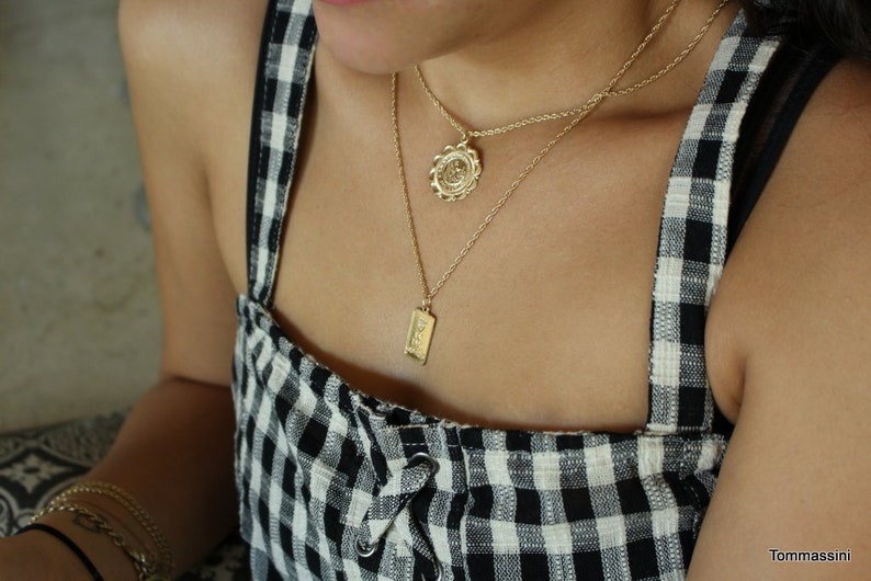 May include: A woman wearing a black and white gingham dress with two gold necklaces. One necklace features a round pendant with a floral design, and the other necklace features a rectangular pendant with an inscription.