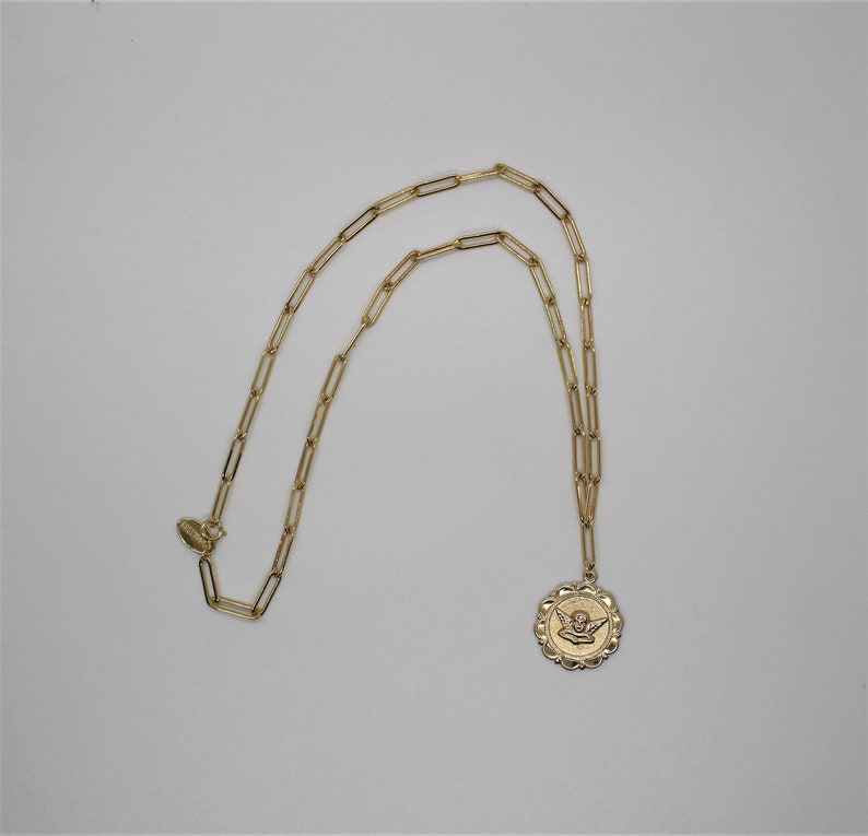 May include: A gold chain necklace with a gold pendant featuring an angel with wings. The pendant is round and has a textured edge.
