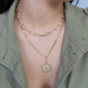 May include: A gold chain necklace with a gold pendant featuring an angel design. The necklace is layered on top of a second gold chain.