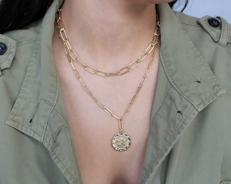 May include: A gold chain necklace with a gold pendant featuring an angel design. The necklace is layered on top of a second gold chain.