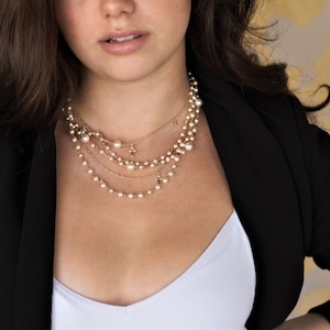 May include: A woman wearing a black blazer and a white tank top with a layered pearl necklace with small gold cross charms.