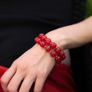May include: A double-strand bracelet featuring large, round, red beads. The bracelet is worn on a wrist, with a black garment and red fabric in the background. The beads are a vibrant red color, and the bracelet appears to be made of polished stones.