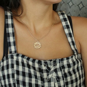 May include: A gold chain necklace with a round pendant featuring a floral design. The necklace is worn by a person wearing a black and white gingham top.