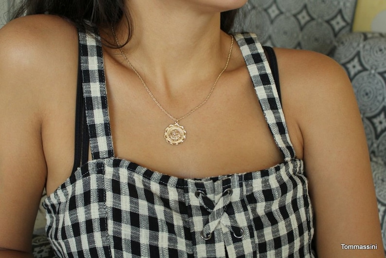 May include: A gold chain necklace with a round pendant featuring a floral design. The necklace is worn by a person wearing a black and white gingham top.
