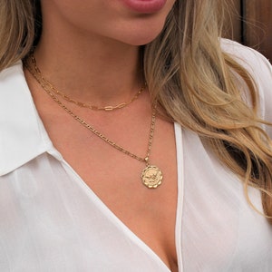 May include: A gold chain necklace with a gold pendant featuring a winged heart design. The necklace is layered over a thinner gold chain.